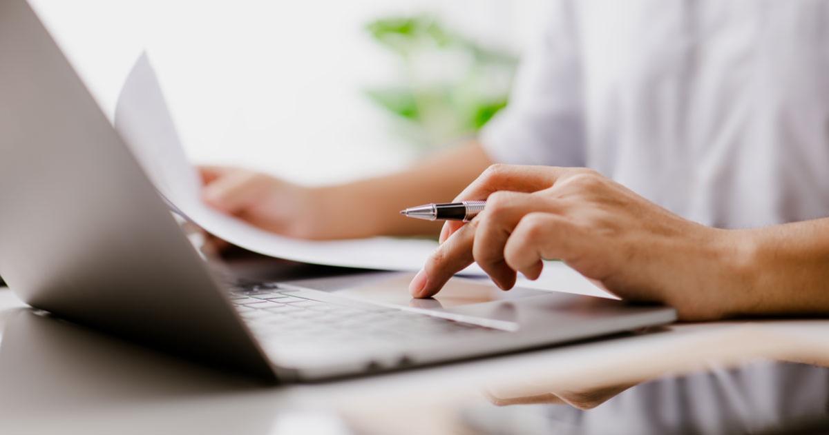 Person sitting at a laptop with a pen and paper in hand