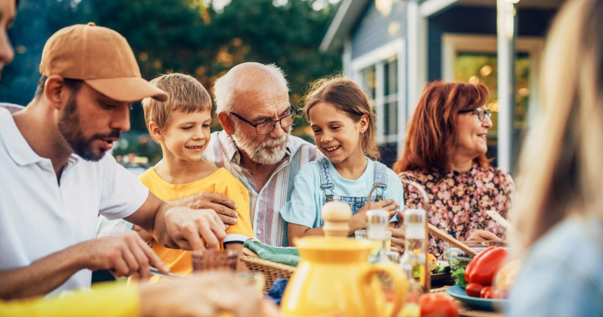 Multigenerational family at a dinner table outside a house, with a grandfather and two grandchildren on his lap at the center