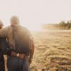The backs of an older man and woman with arms around each other hiking into the sunset.