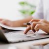 Person sitting at a laptop with a pen and paper in hand