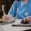 Person in scrubs with stethoscope using a calculator, notebook and laptop